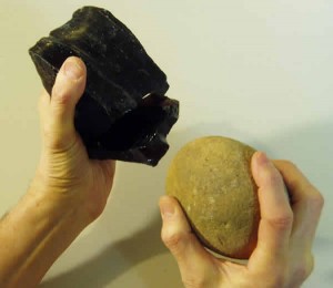 Hands hold a hammer stone and chunk of obsidian